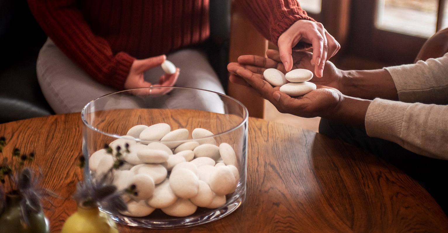 Two people exchanging white pebbles over a glass bowl filled with pebbles on a wooden table