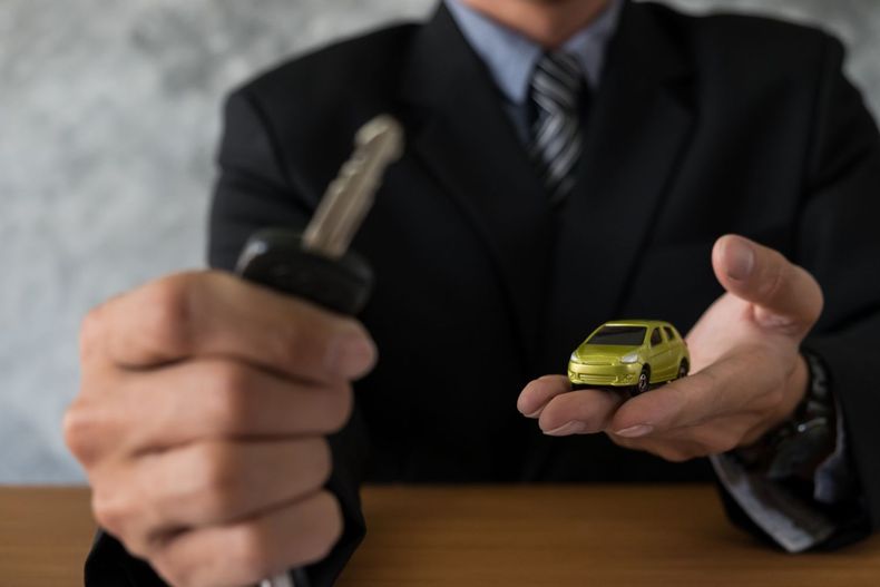 Una persona in giacca e cravatta tiene in mano una chiave dell'auto vicino a un piccolo modellino di auto gialla su un tavolo.
