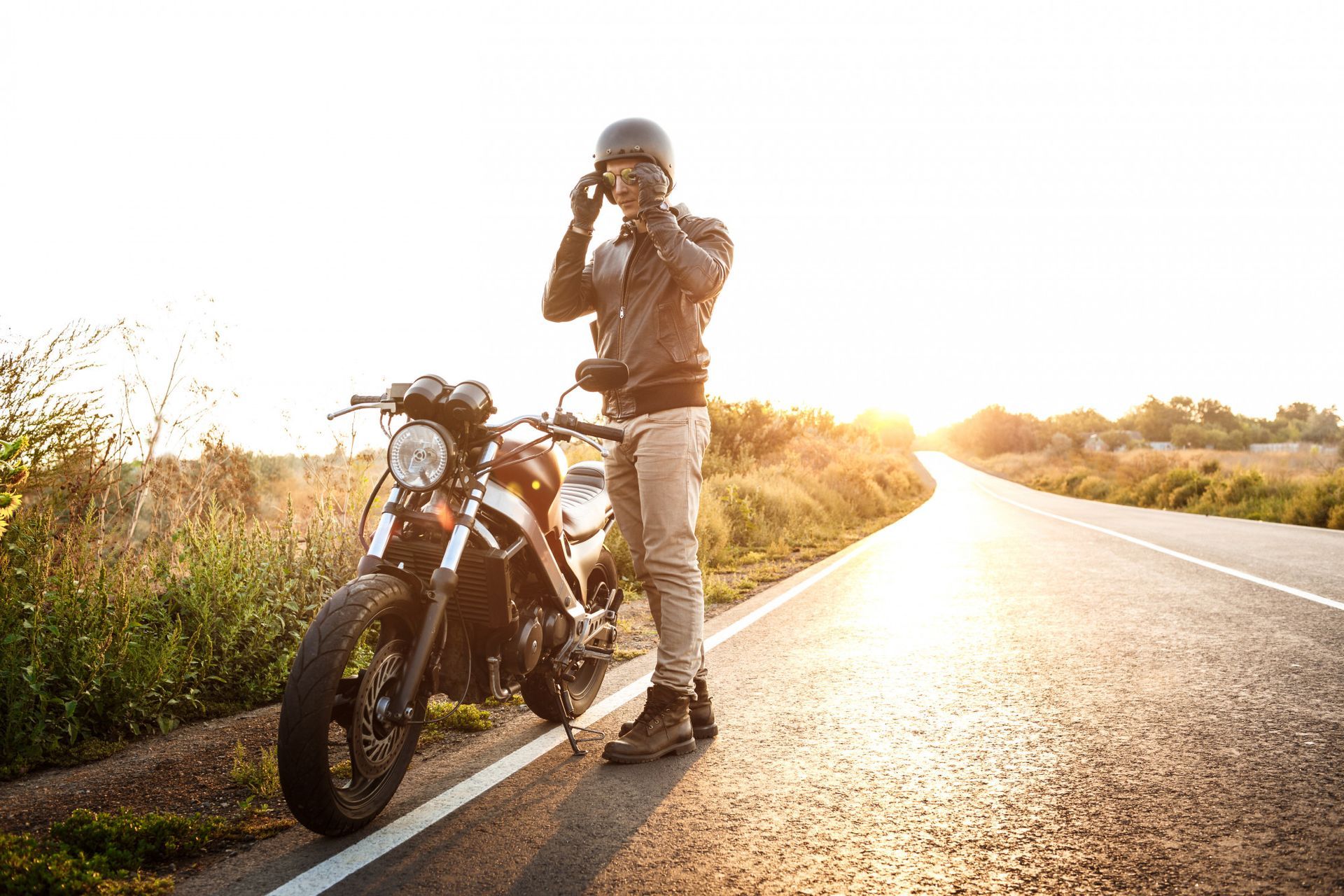 Un motociclista in piedi accanto a una moto nera sul ciglio della strada illuminato dal sole al tramonto.