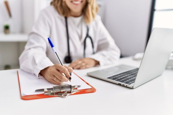 Un medico prende appunti su un blocco per appunti accanto a un computer portatile in un ufficio luminoso.