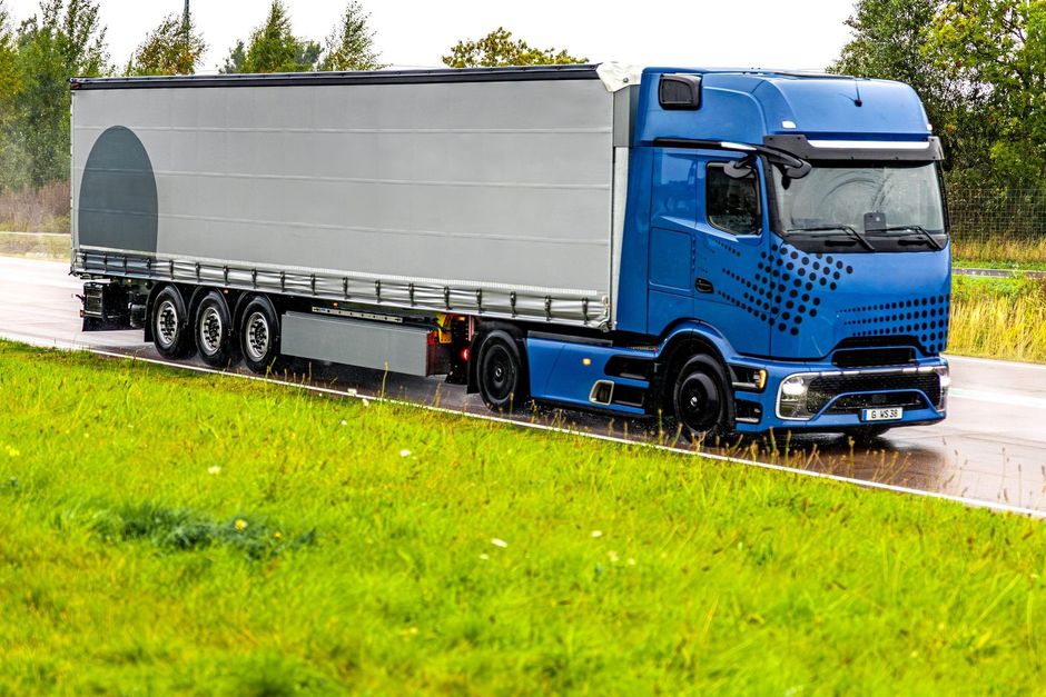 Un camion blu con rimorchio grigio percorre una strada bagnata accanto a un campo erboso.