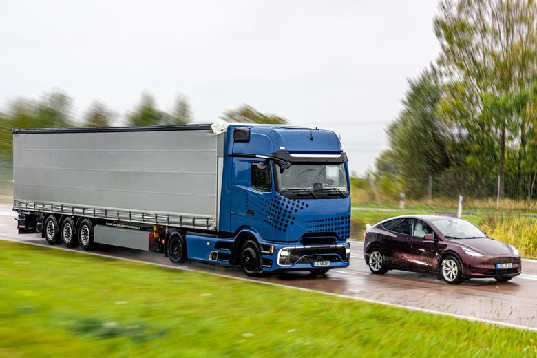 Un camion blu che traina un'auto bordeaux su una strada bagnata accanto a campi erbosi.