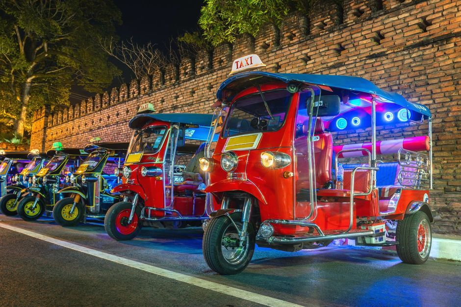 Una fila di coloratissimi tuk-tuk rossi e blu parcheggiati di notte lungo un muro illuminato della strada.