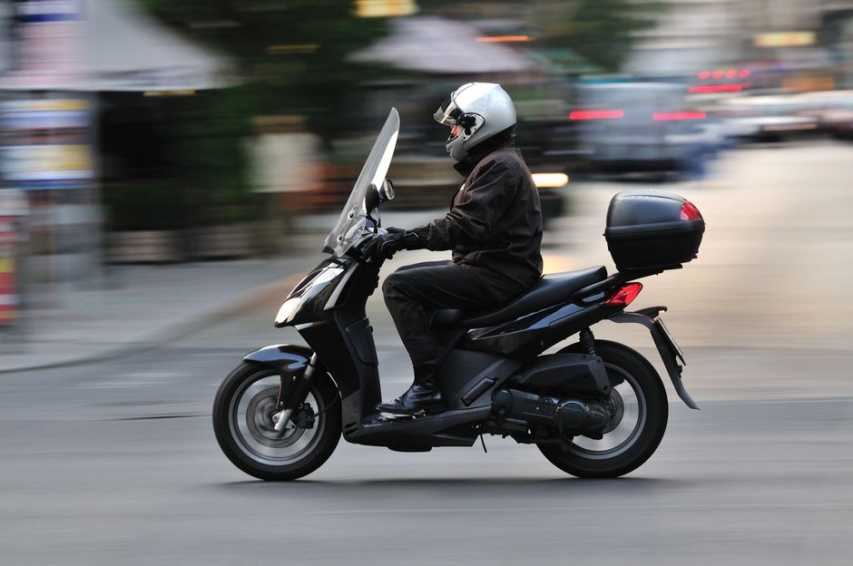 Un motociclista con un casco bianco alla guida di uno scooter nero percorre una strada cittadina sfocata.