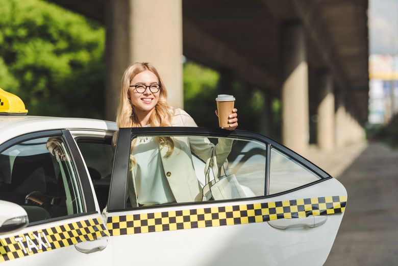 Una donna sorridente è in piedi accanto a un taxi, con in mano una tazza di caffè, in una strada cittadina.