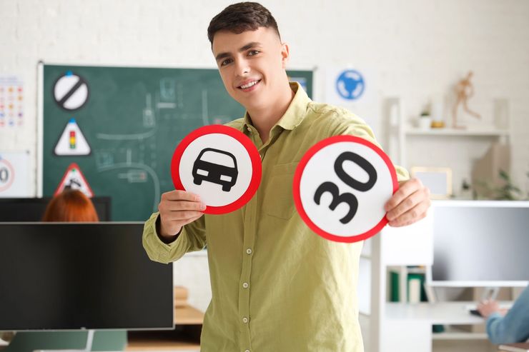 Un uomo tiene in mano due cartelli stradali con bordo rosso in un'aula scolastica: divieto di sorpasso e limite di velocità 30.