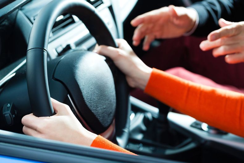 Mani sul volante di un'auto, con un passeggero che gesticola accanto a loro.