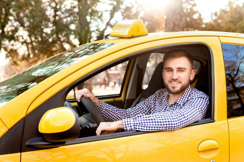 Autista sorridente in un taxi giallo, seduto al volante con la luce del sole sullo sfondo.