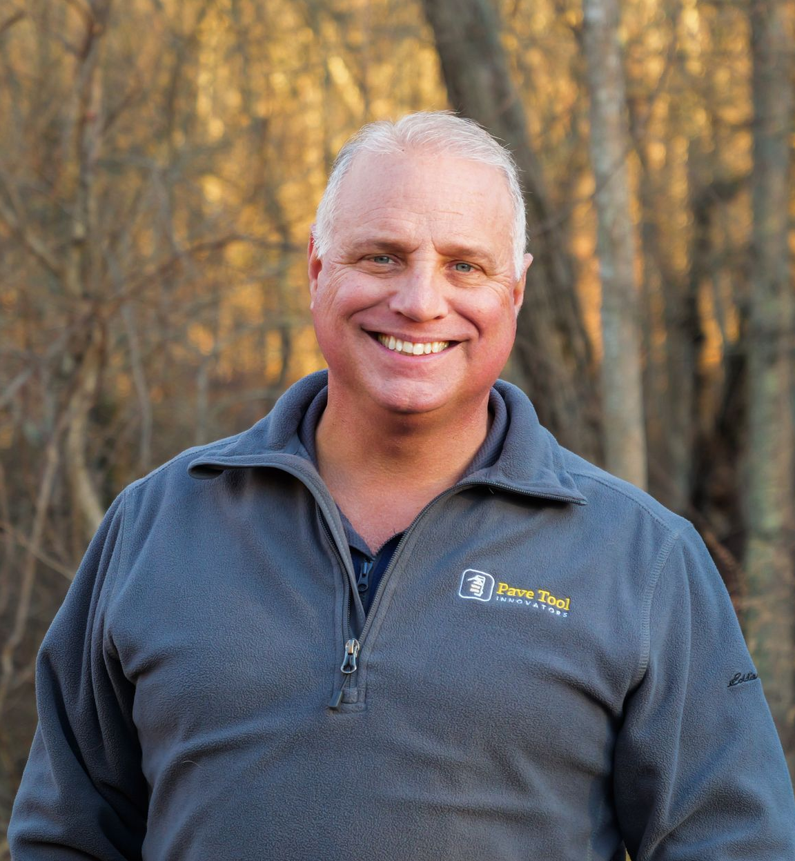Man smiling in a blue and gray jacket with a company logo in front of a wooden background.