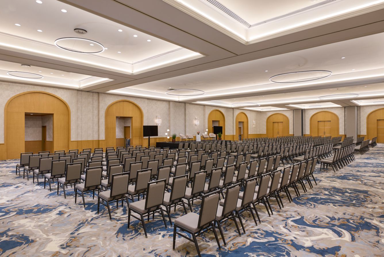 Empty auditorium with rows of tan chairs, blue carpet, and decorative ceiling lights.