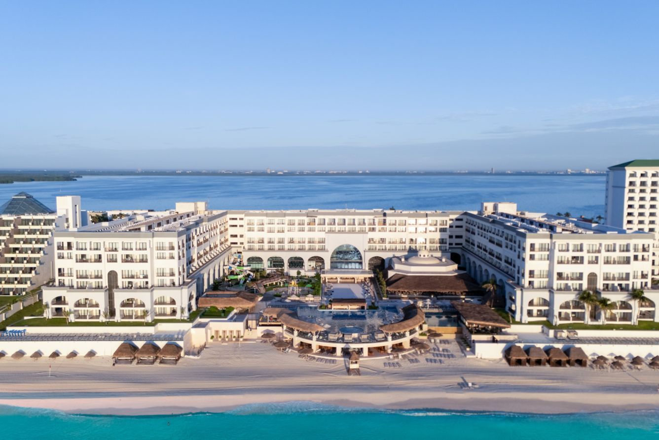 Bright blue ocean and sky over a beach with palm trees and a resort, under a partly cloudy sky.