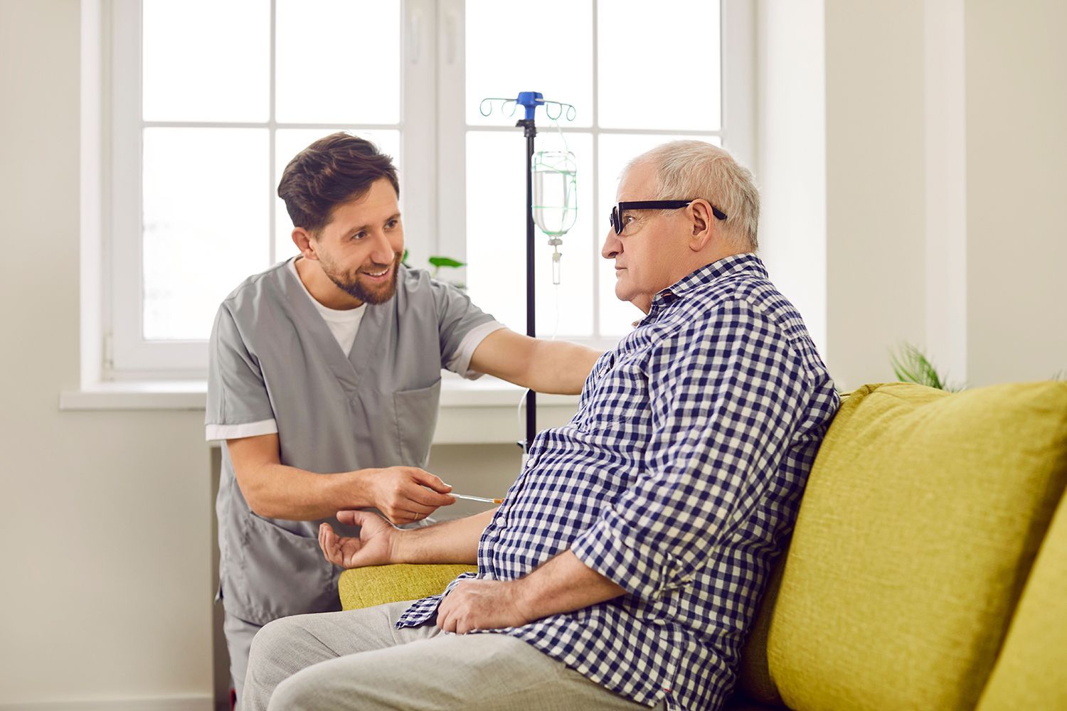 Healthcare worker talking with a seated patient in a bright living room, beside a green couch and IV stand