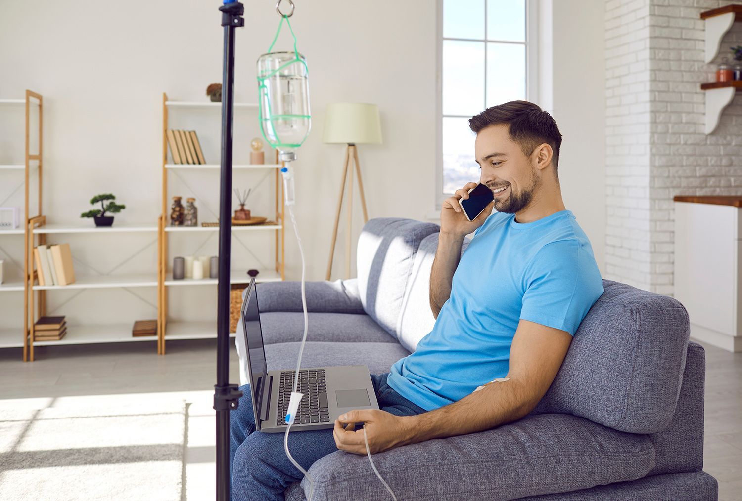 Man sitting on a gray sofa, speaking on a phone beside an IV drip in a bright living room.