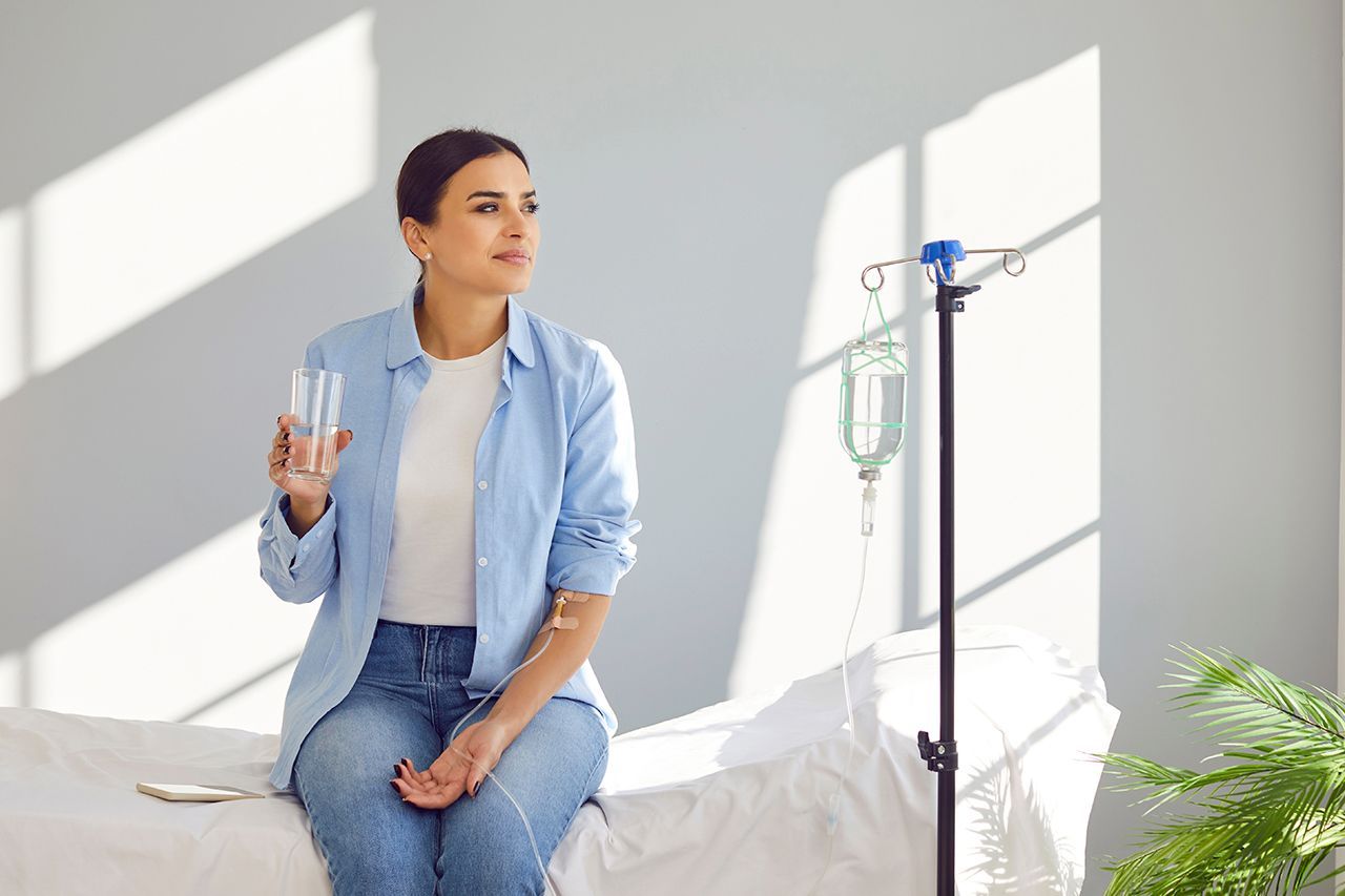 Woman in a blue shirt sits by an IV stand, holding a glass of water in a bright room.