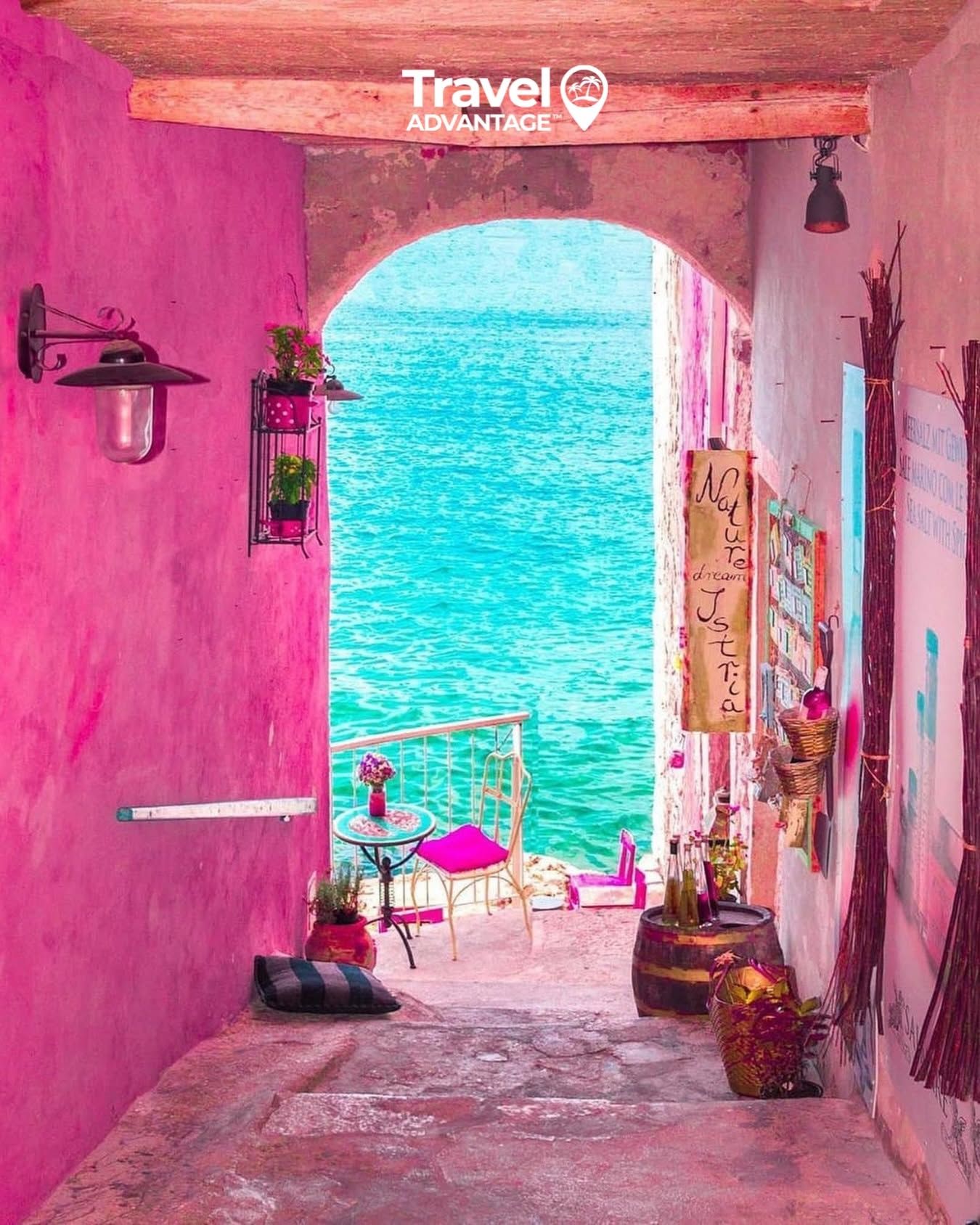 Pink alley opening to bright turquoise sea, with a small chair and potted plants by the water.