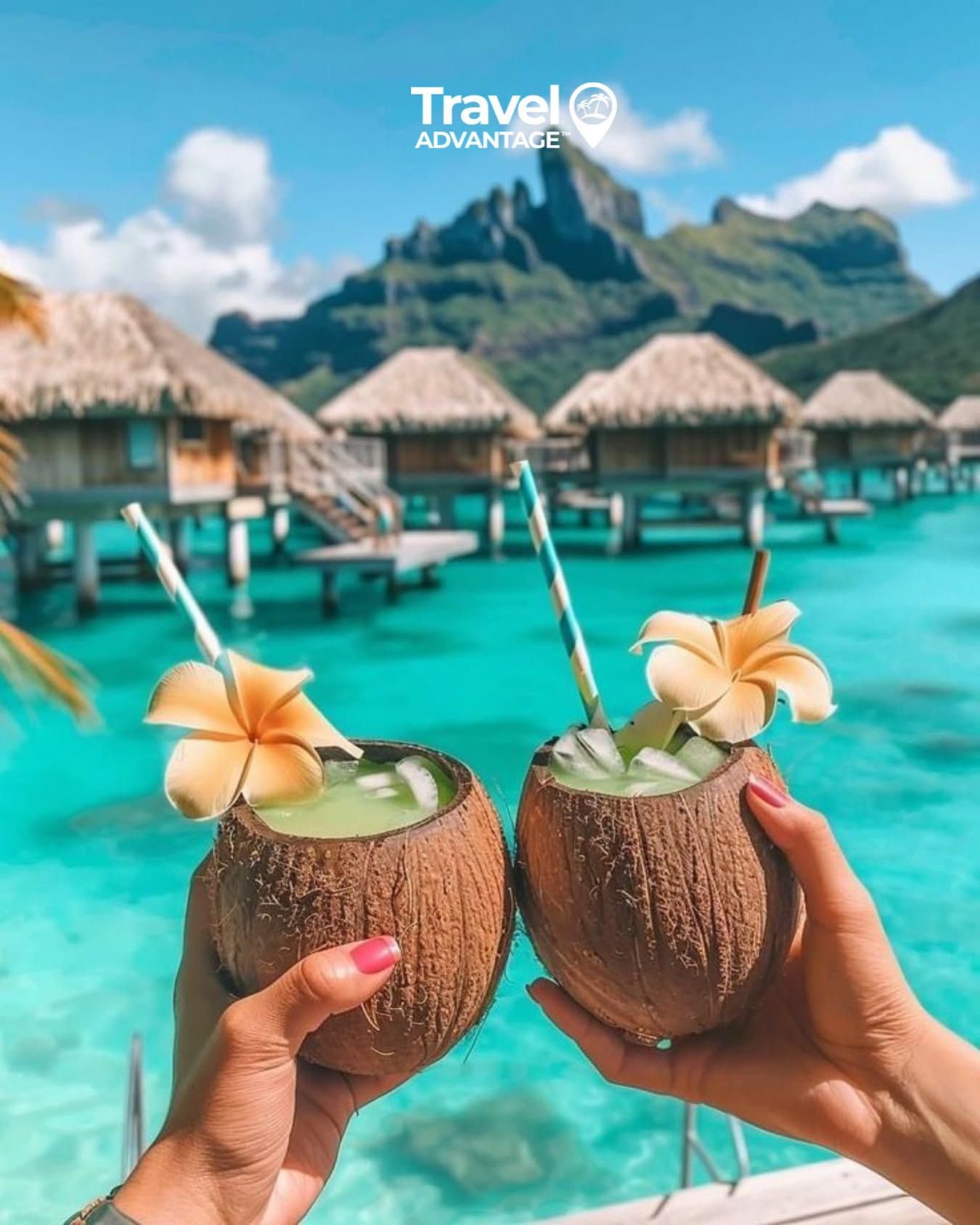 Two hands holding coconut drinks with flowers over turquoise overwater bungalows and mountains.