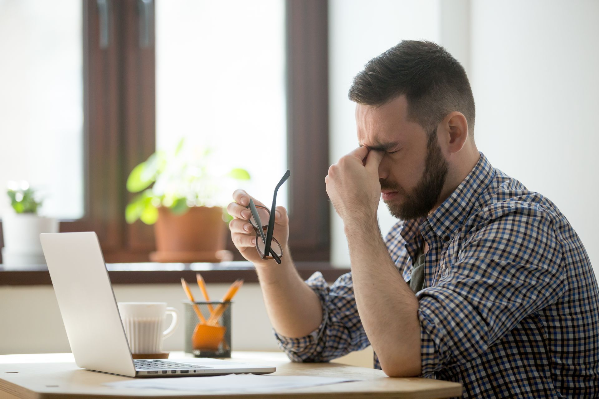 A man is sitting at a desk with a laptop and holding his glasses.