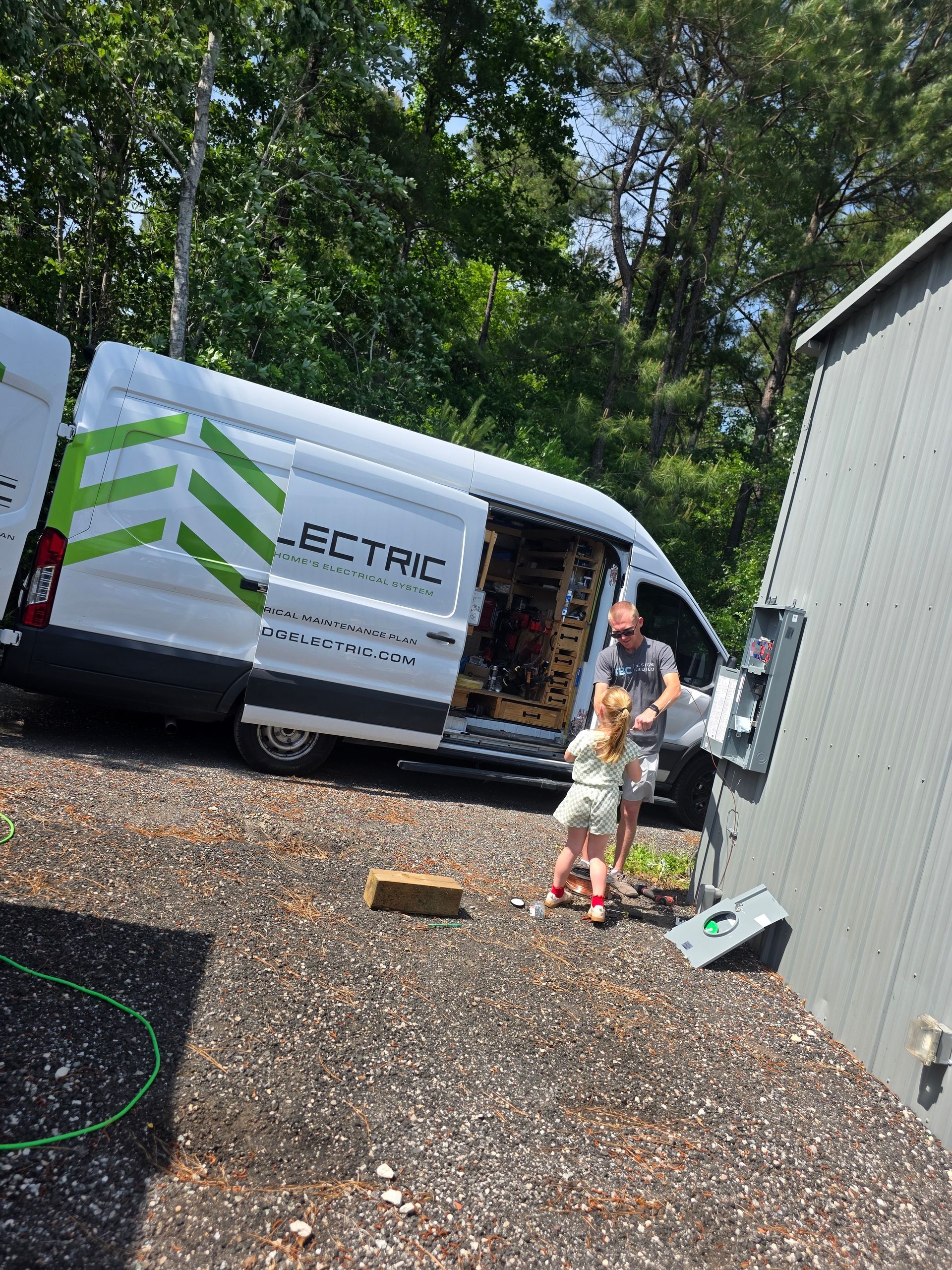 Person and child beside white electric service van in a gravel lot, with tools and a metal shed nearby.