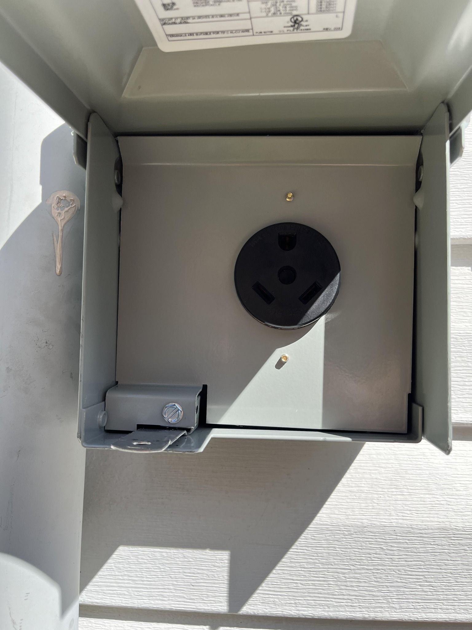 Outdoor electrical outlet box with a black, three-prong receptacle; the box is open on a grey wall.