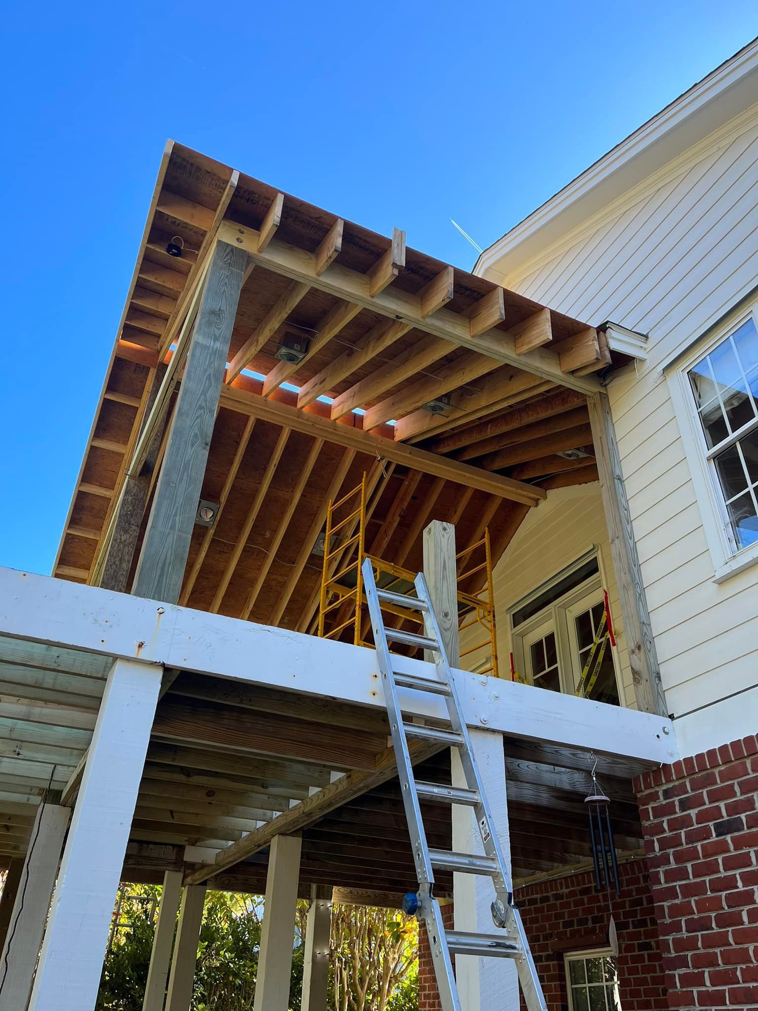 Construction of a wooden porch roof attached to a white house with a brick foundation, a ladder, and a blue sky.