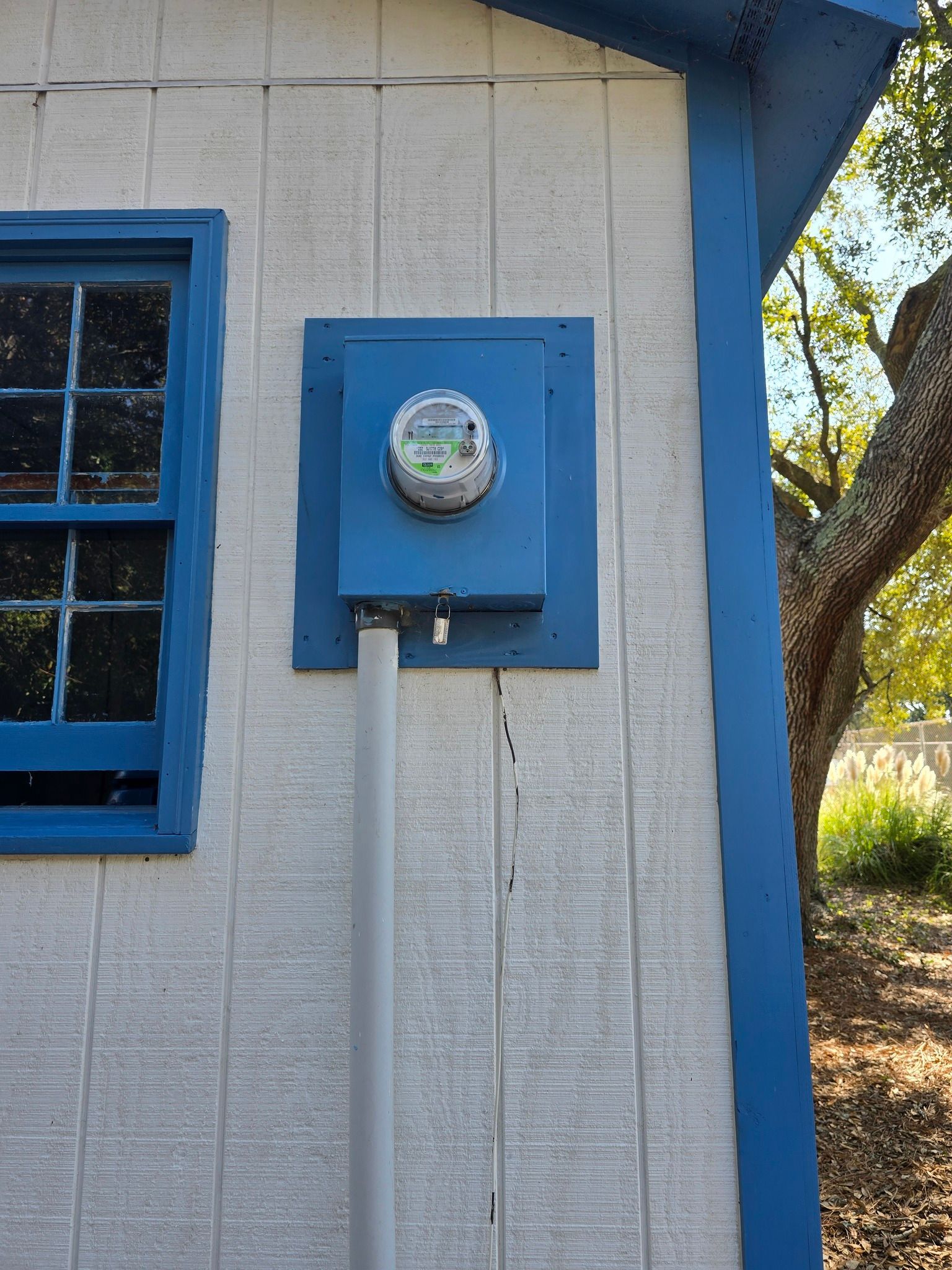 Exterior wall with a blue electric meter box and conduit. White siding with blue trim and a window.
