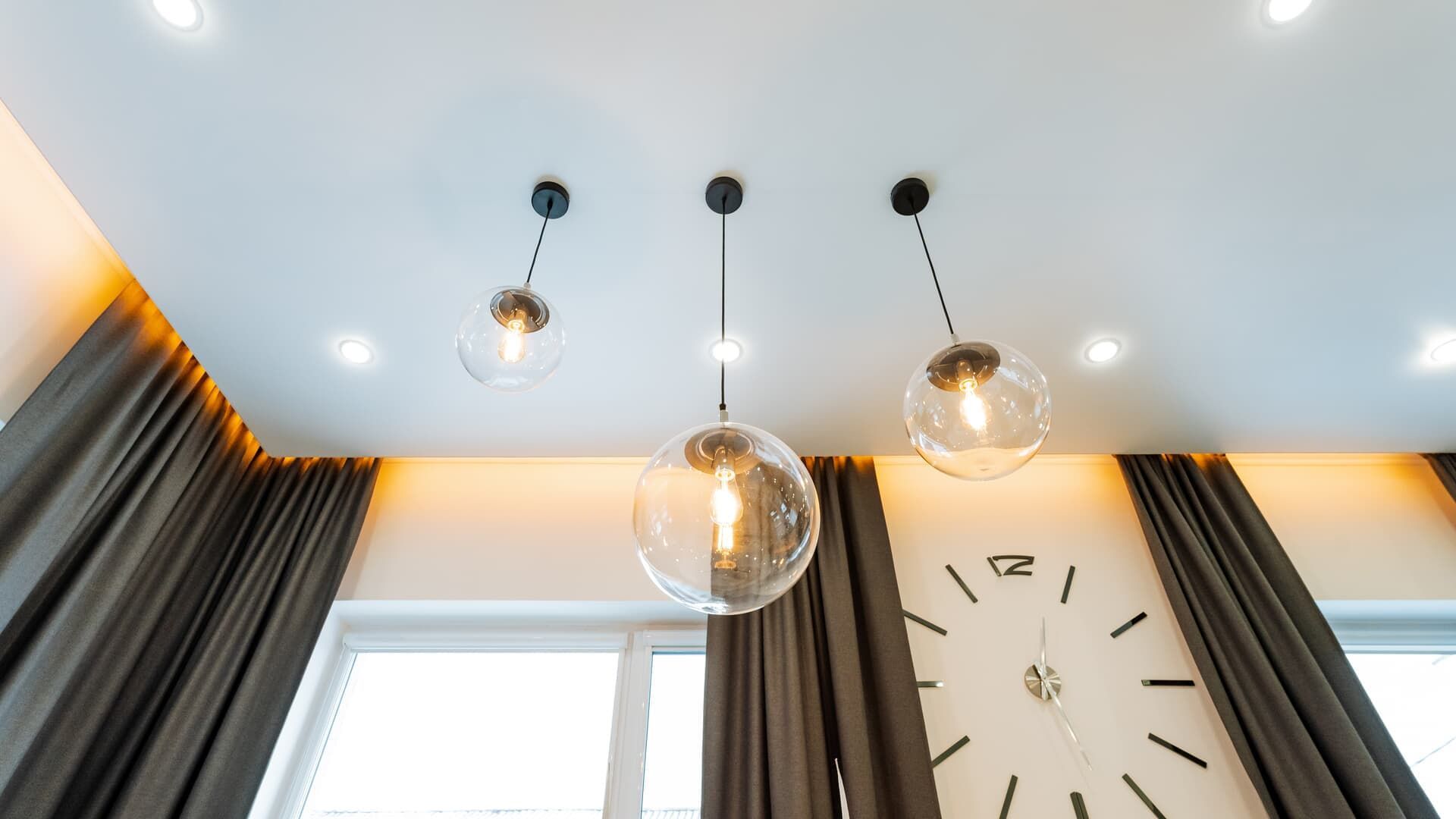 Three globe pendant lights hanging from a white ceiling, next to a large clock and curtains.