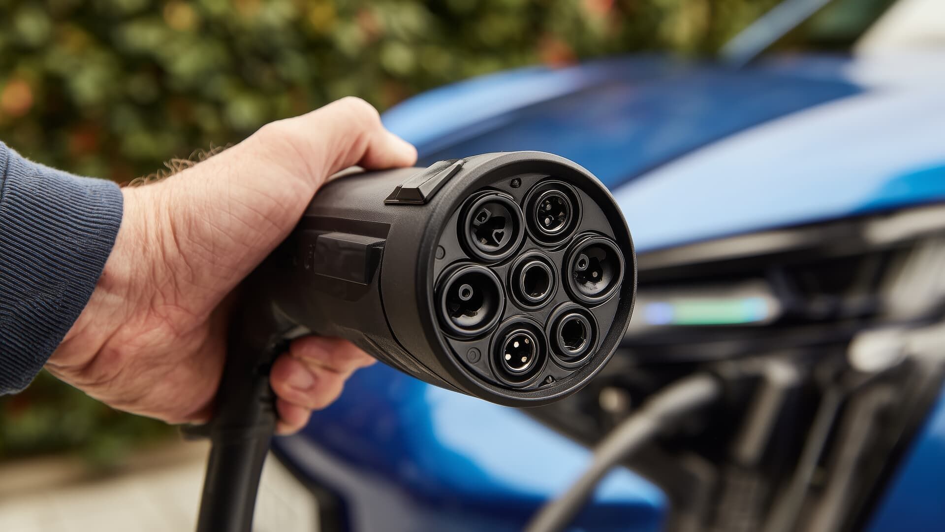 Hand holding an EV charging cable, connecting to a blue car.