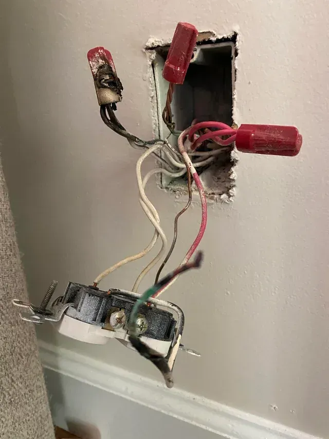 Exposed electrical wiring in a wall opening with red wire nuts and a loose outlet hanging below.