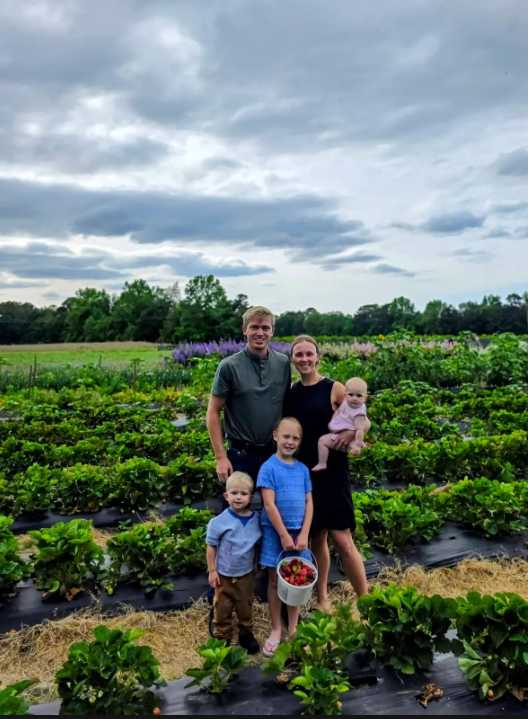 Family posing in a green field of plants under a cloudy sky, holding a basket of fresh vegetables.