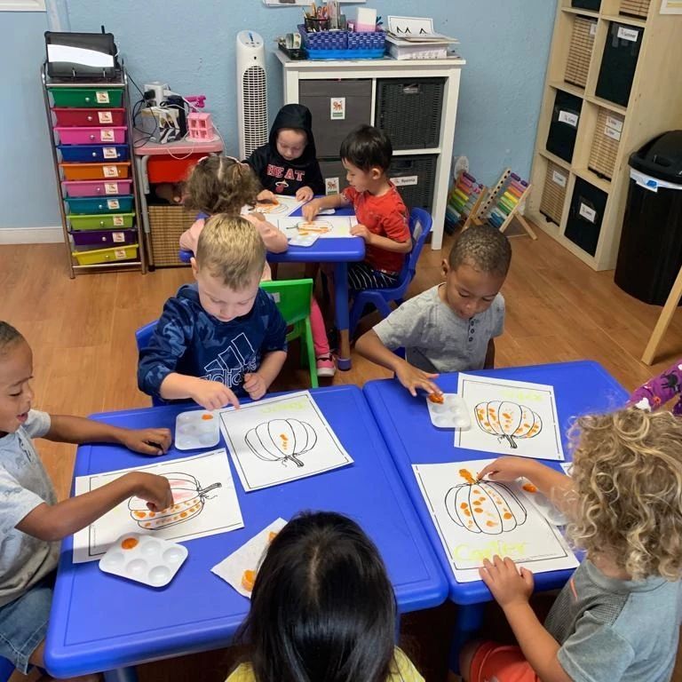 A group of children are sitting at blue tables drawing pumpkins