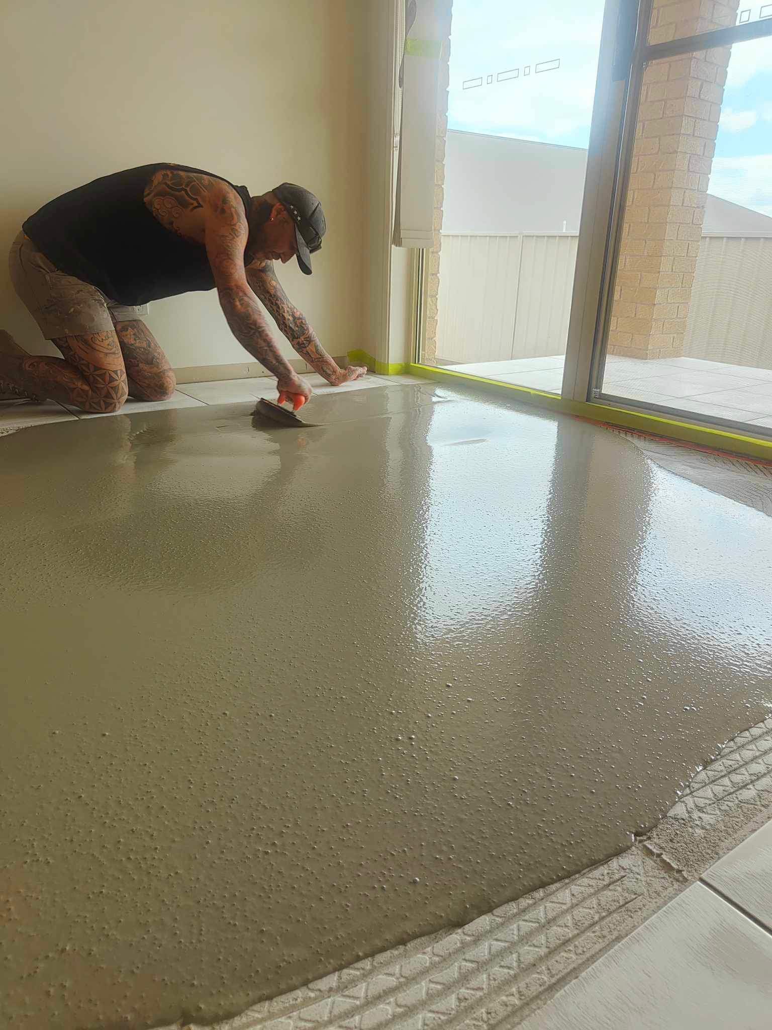Man kneeling, smoothing wet concrete floor. Interior room, sunlight, tan/gray tones.