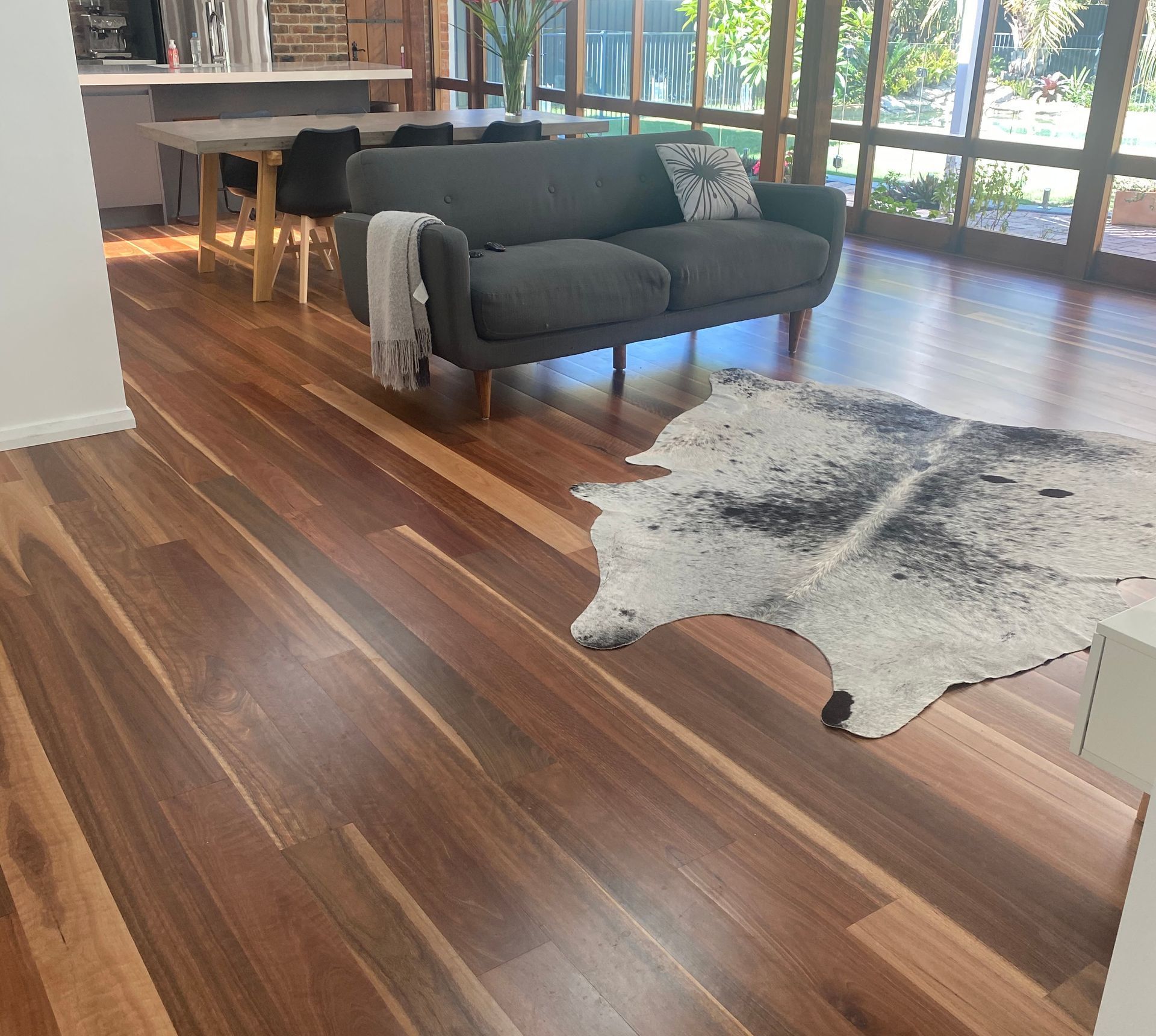 Wooden floor with dark gray couch, cowhide rug, and dining area in a sunlit room.