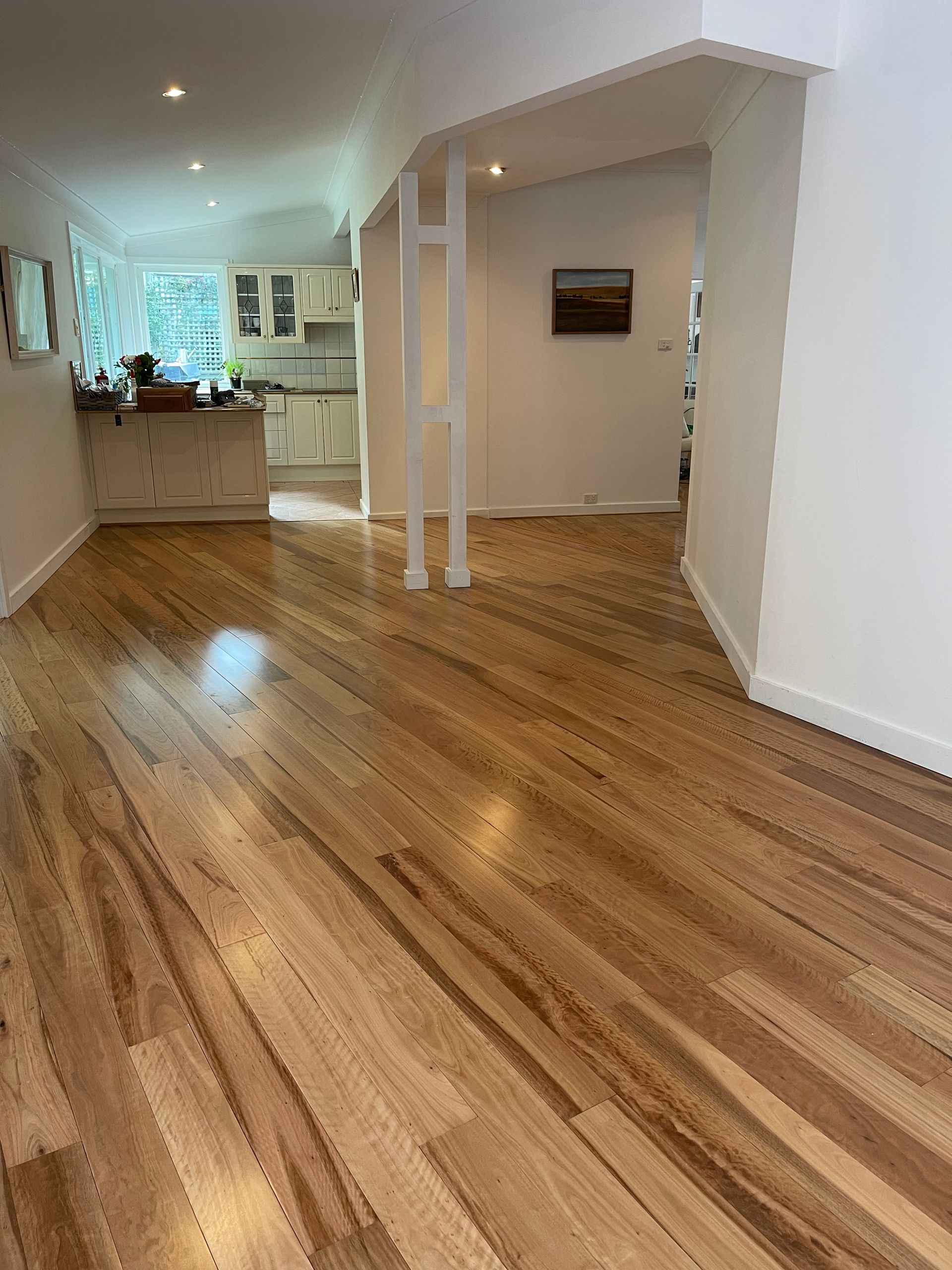 Hardwood floors in a light-filled room, leading to a kitchen with white cabinets, and a hallway with artwork.