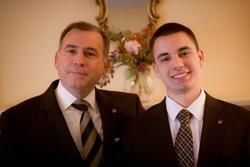 Two men in suits smiling indoors, standing before a floral arrangement.