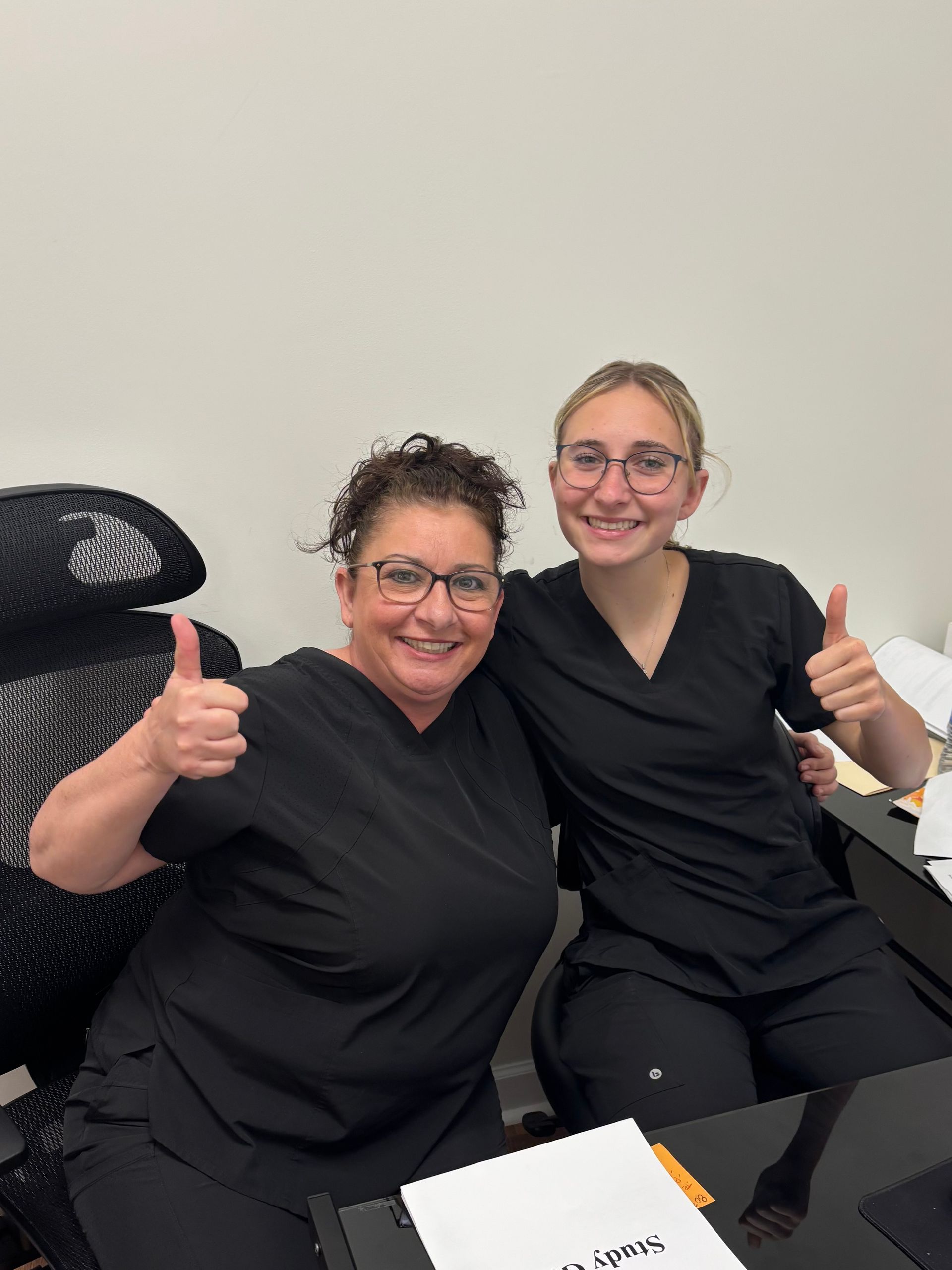 Two women in black scrubs giving thumbs up, smiling in an office setting.