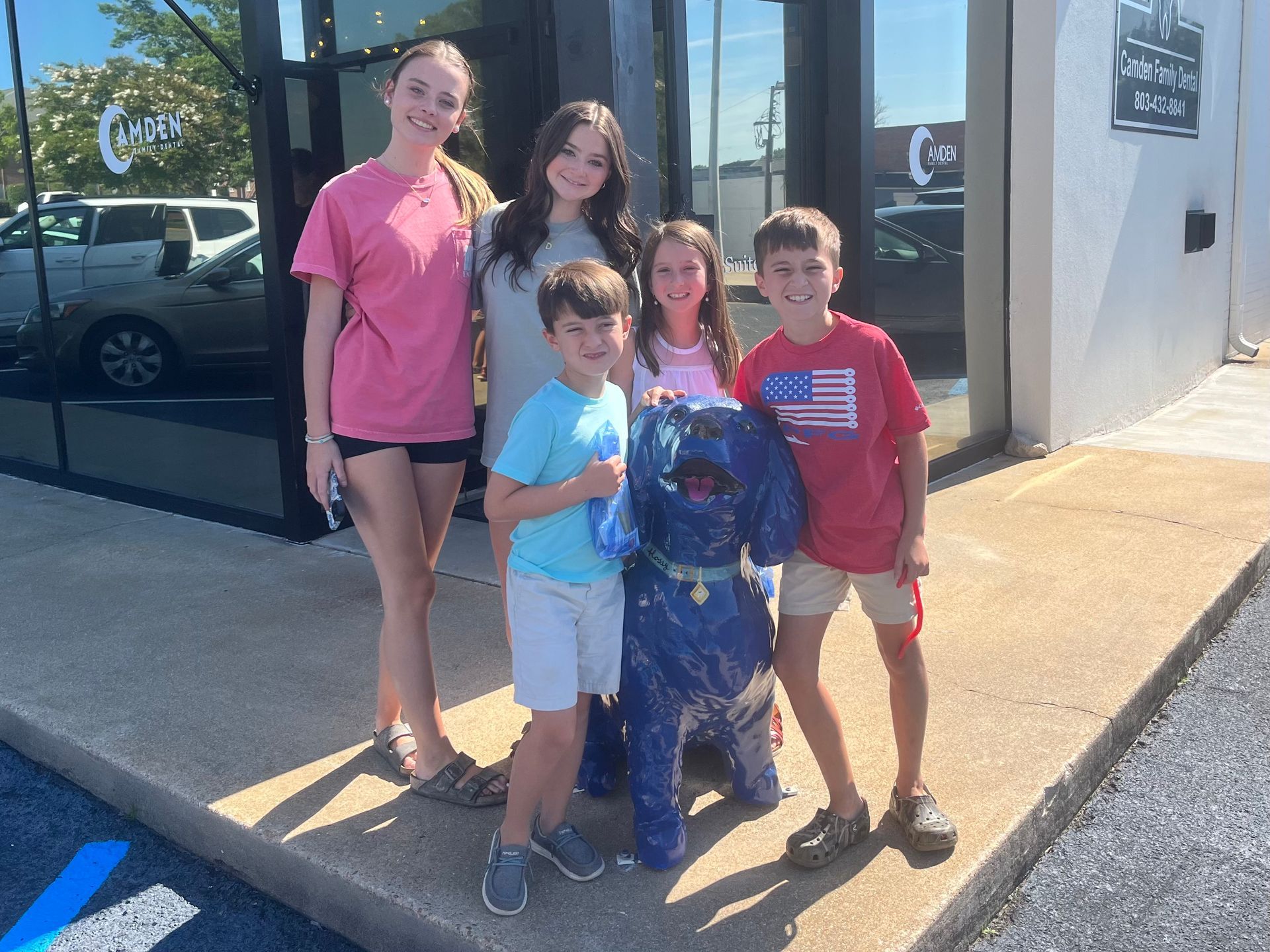 Five kids pose with a blue dog sculpture in front of a building with a sign.