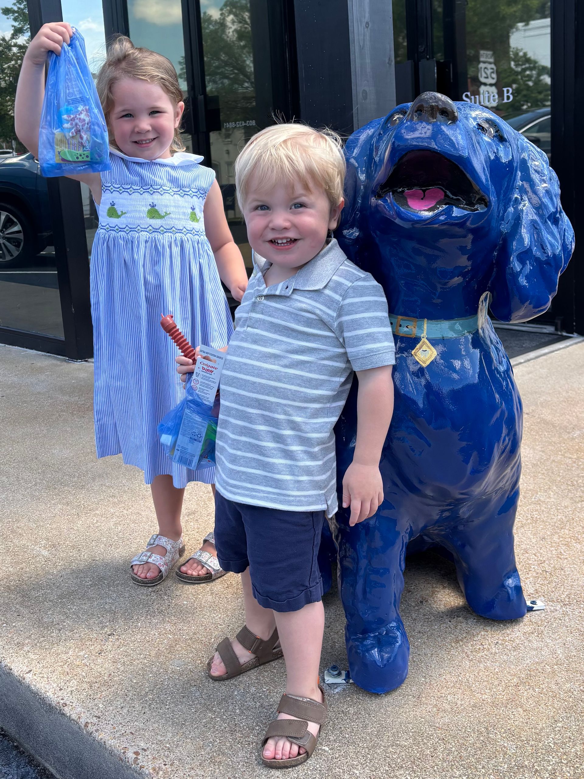 Two young children smiling with a blue dog statue outside; one holds a treat bag, the other a lollipop.