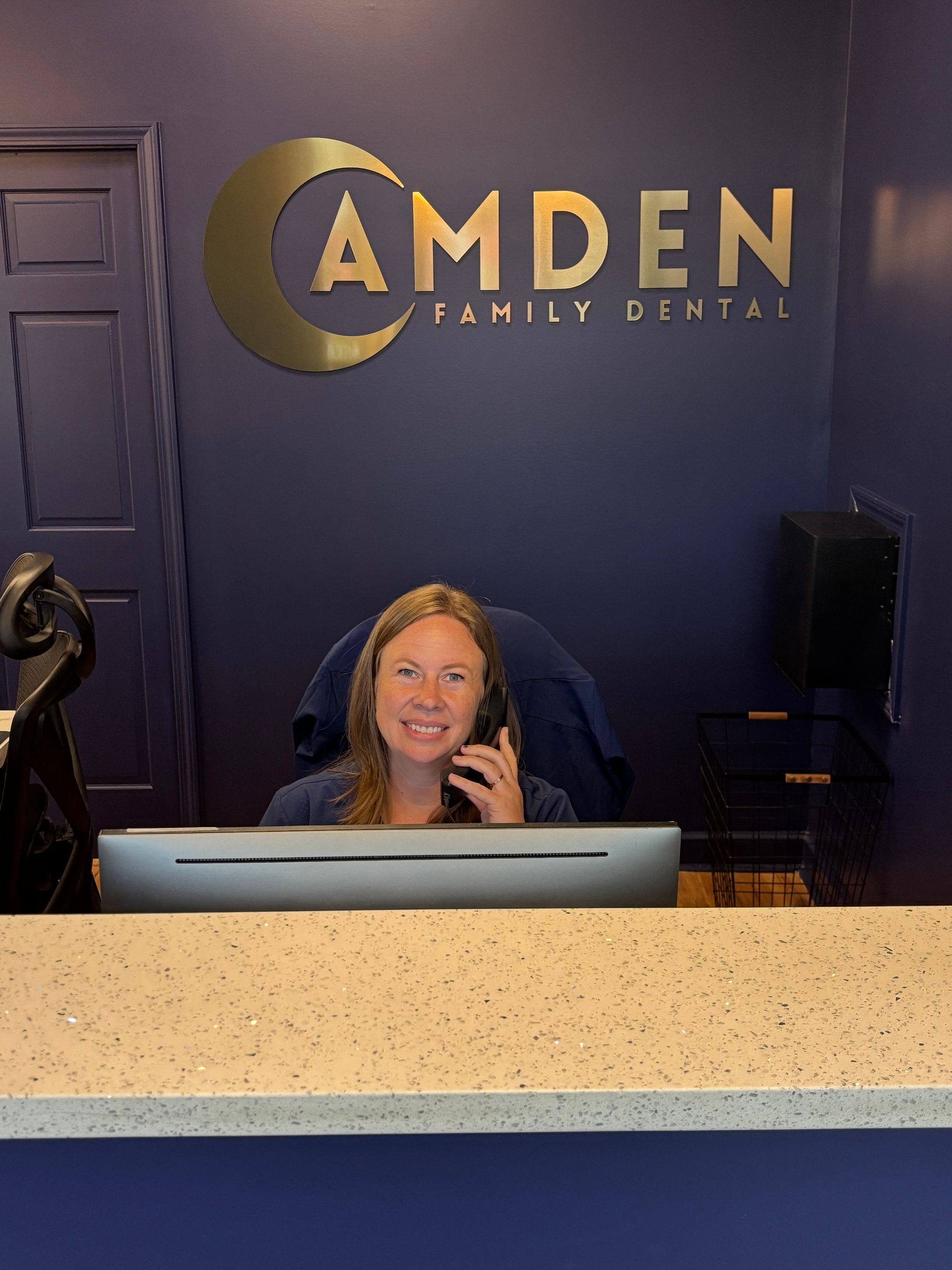 Woman at Camden Family Dental reception desk, smiling, talking on phone, blue wall, gold sign.
