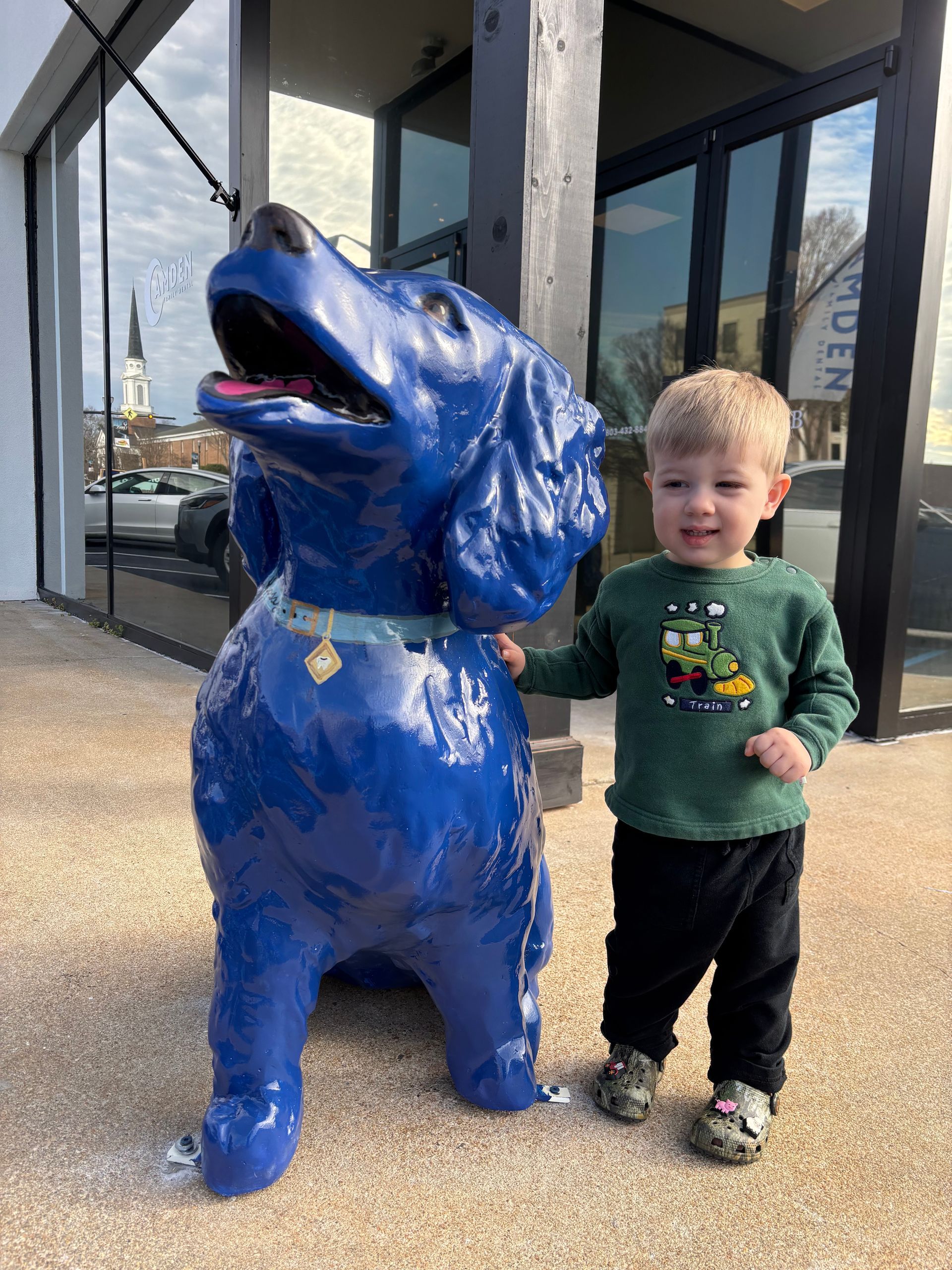 Young child stands next to a large blue dog statue outdoors.