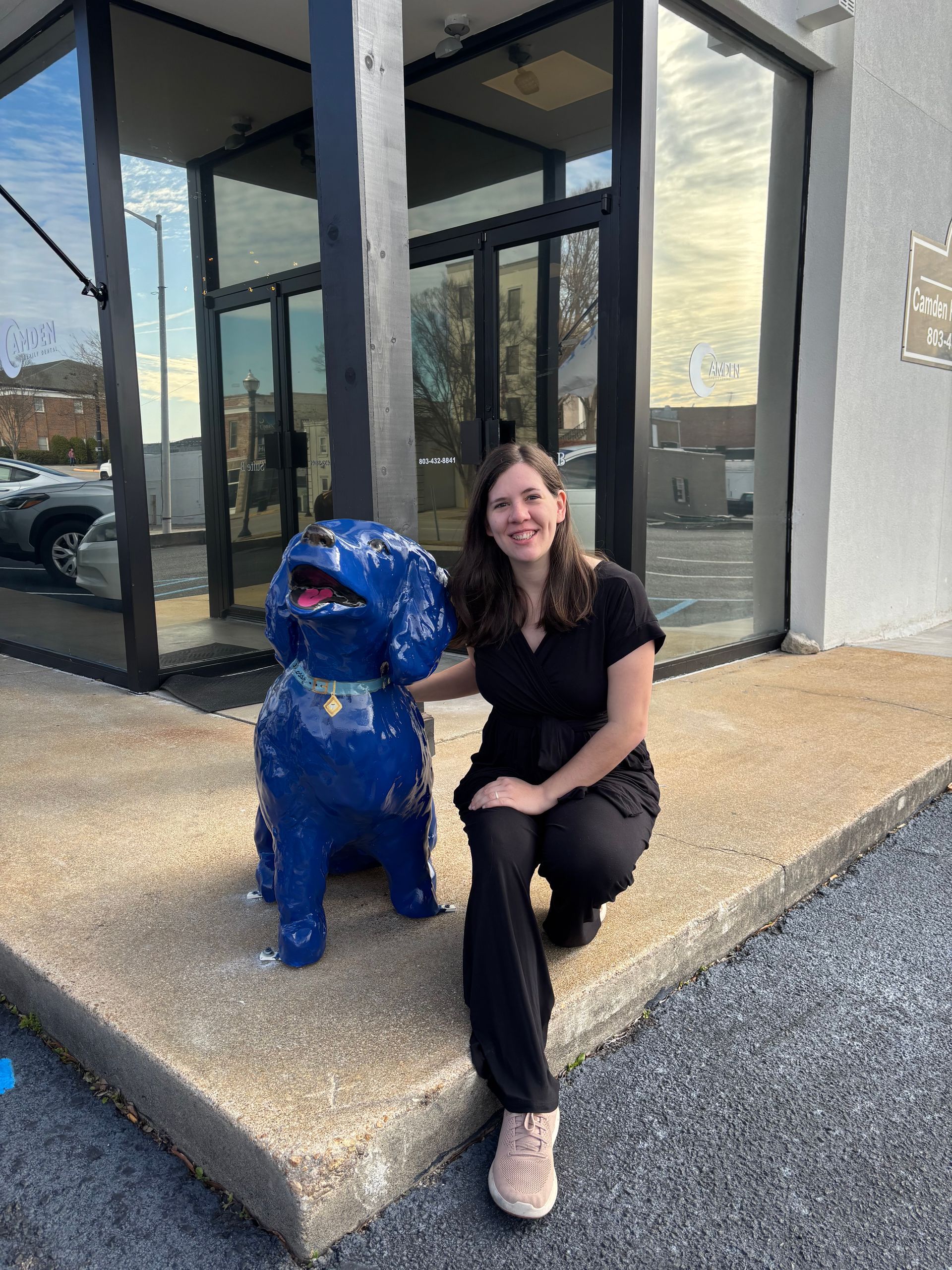 Woman kneels next to a blue dog statue outside a building with glass doors.