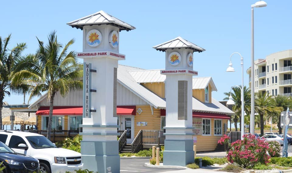 Decorative pillars at a sunny marina entrance with palm trees, parked cars, and a red-roofed building