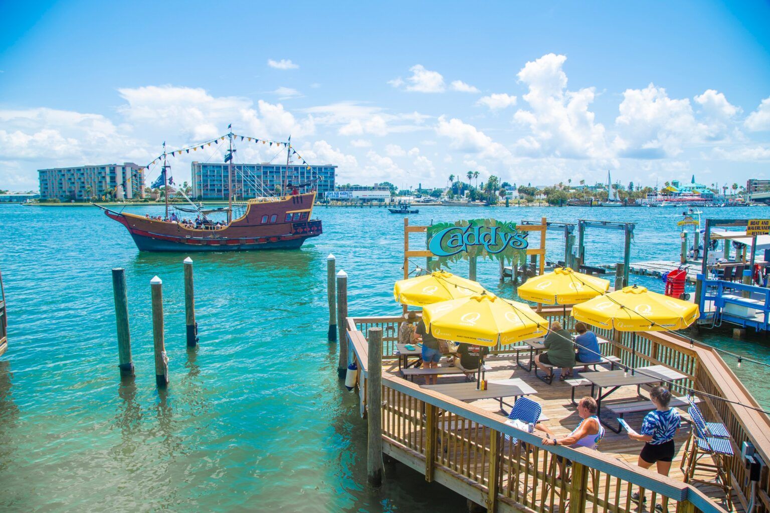 Tropical waterfront deck with yellow umbrellas, people lounging, and a boat on turquoise water under a blue sky