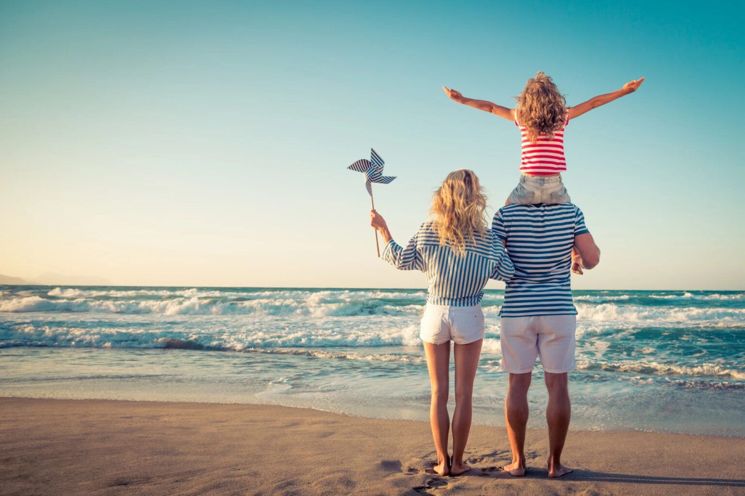 Three people stand on a sandy beach facing the ocean, one child sitting on an adult’s shoulders, arms raised.