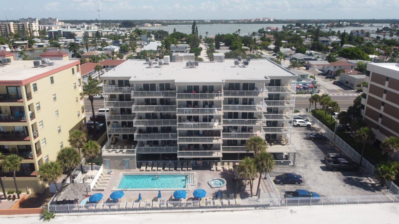 Aerial view of a beachfront condo with a swimming pool and palm trees, near water and neighboring buildings