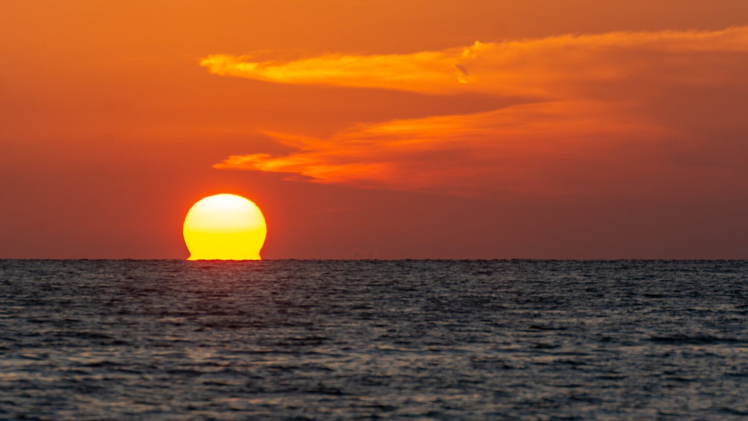 Sun setting over a calm ocean, with orange sky and dark horizon.