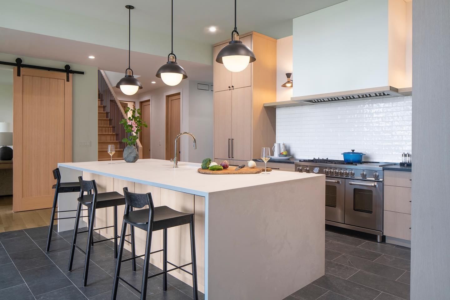Modern kitchen with a large island, black stools, pendant lights, and stainless-steel appliances.