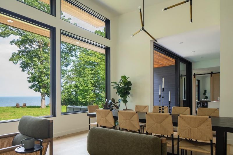Modern dining room with large windows overlooking a green lawn and water.