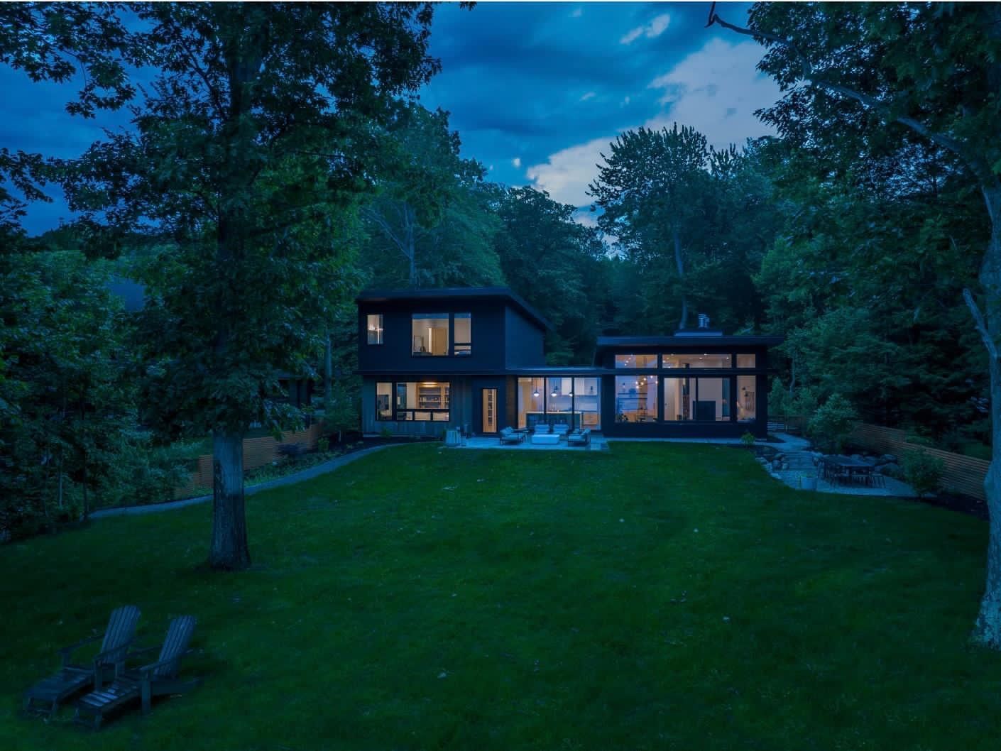 Modern house with lit windows in a wooded backyard at dusk, viewed across a large green lawn.