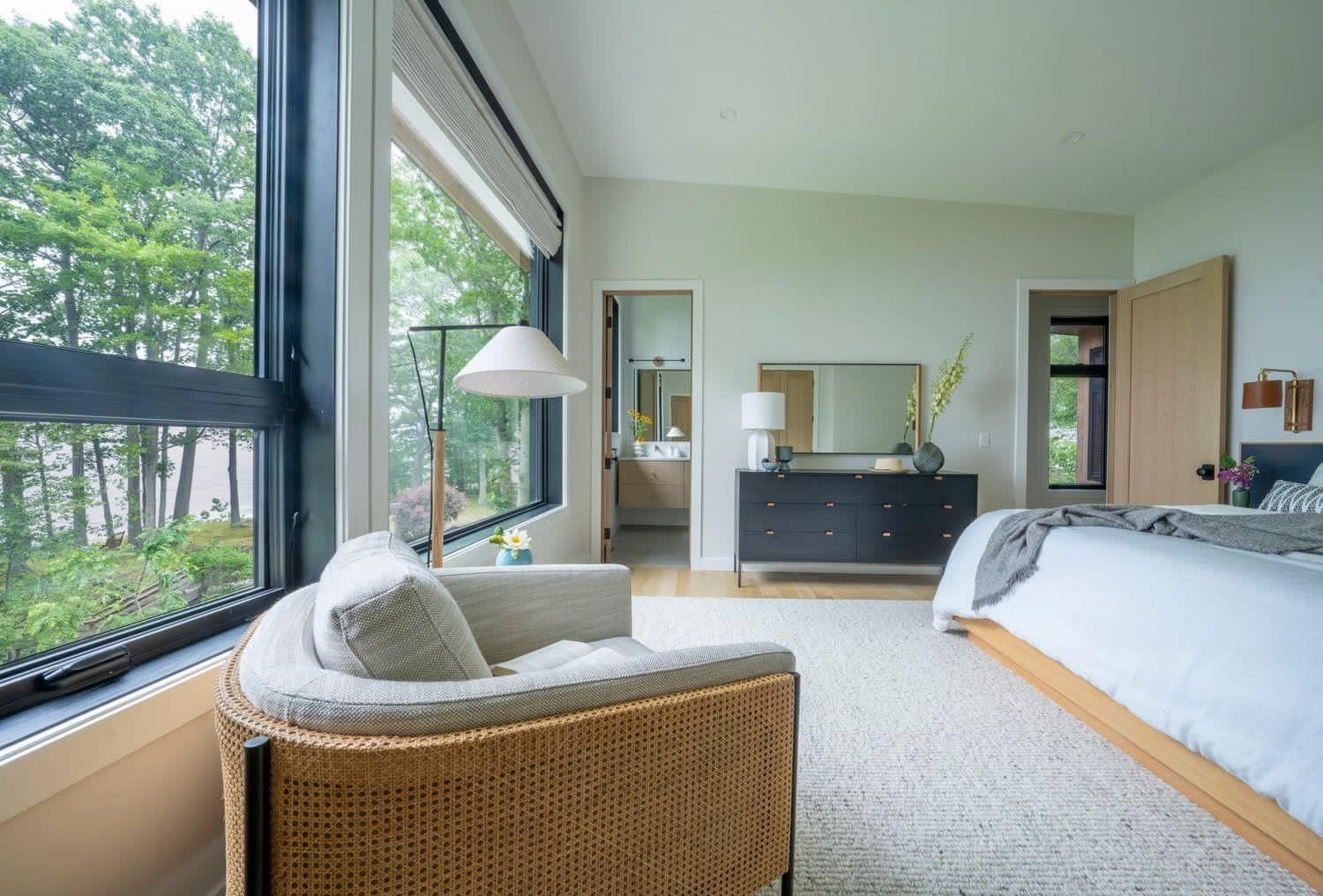 Bright bedroom with large windows, wicker chair, bed, and dresser overlooking green trees