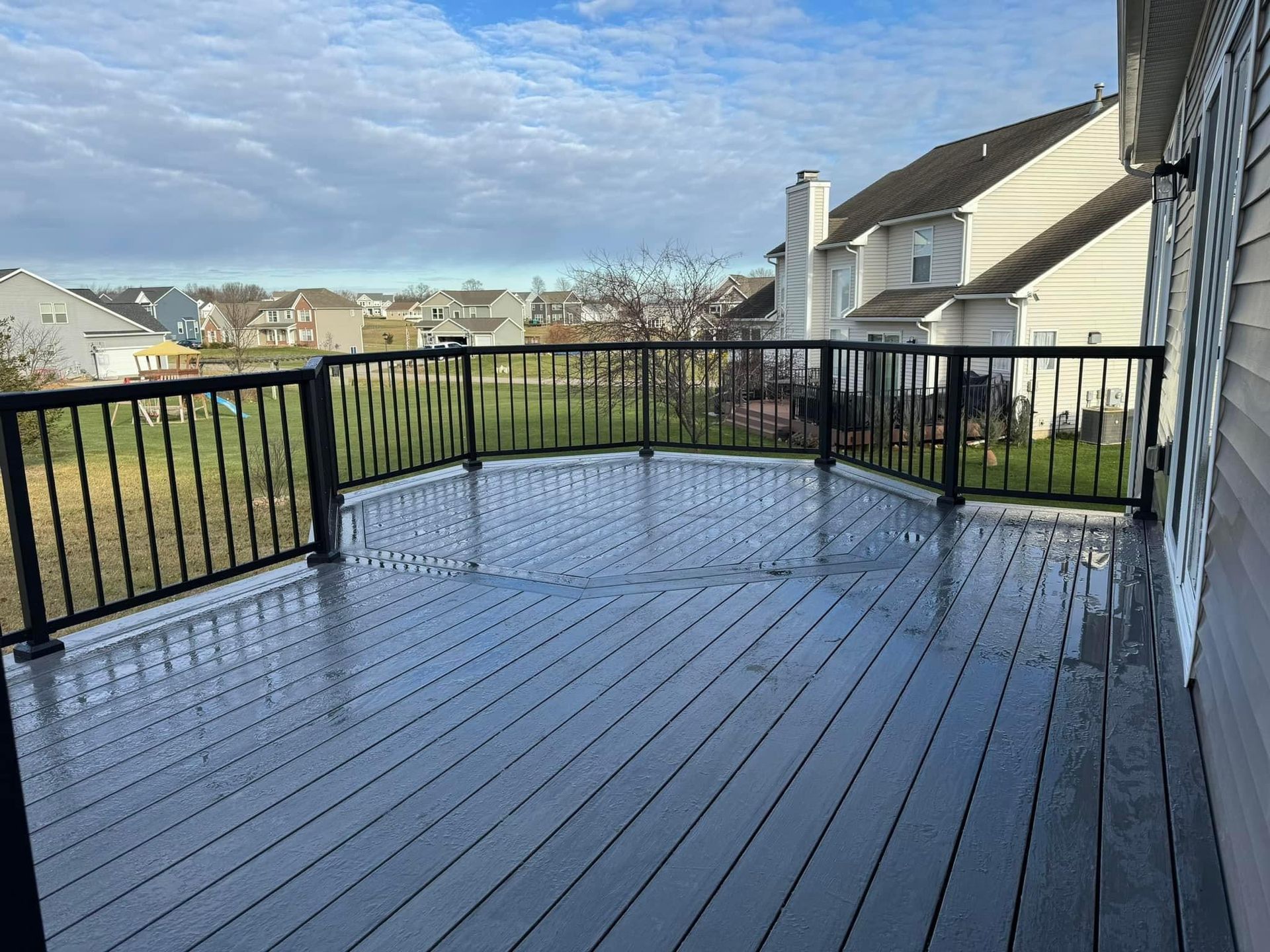 Wet wooden deck with black railing overlooking a grassy neighborhood under a cloudy sky